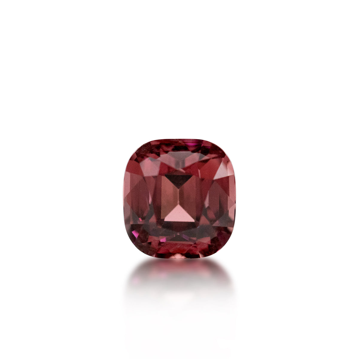 A cushion-cut pinkish-red gemstone with a faceted cushion shape, centered on a clean white background with a soft reflection below.