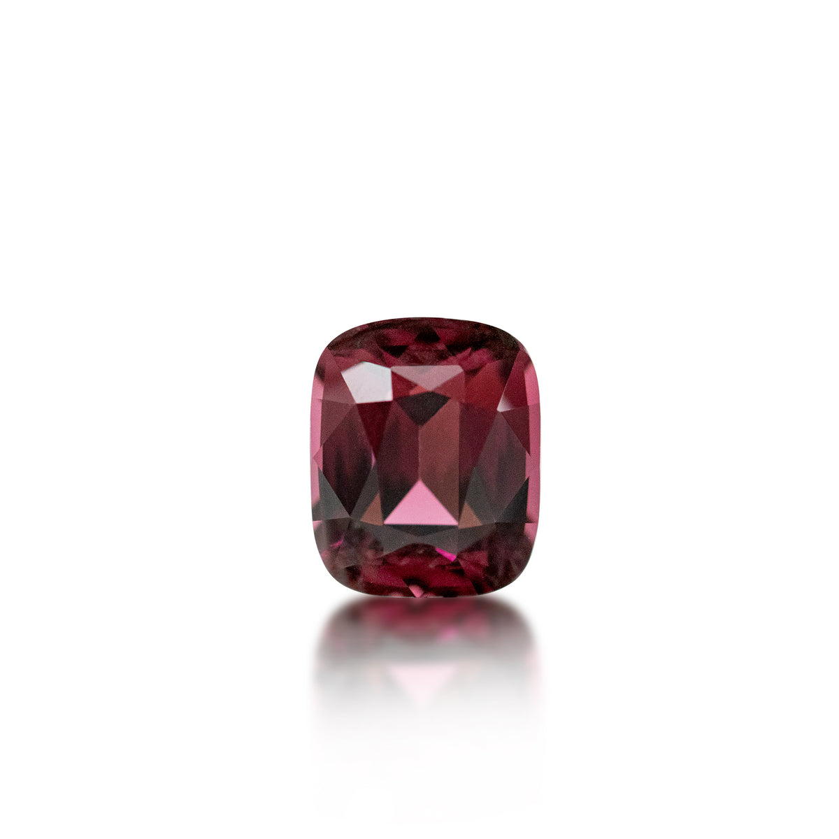 A cushion-cut pinkish-red gemstone with faceted geometric cuts, centered on a clean white background with a soft reflection below.