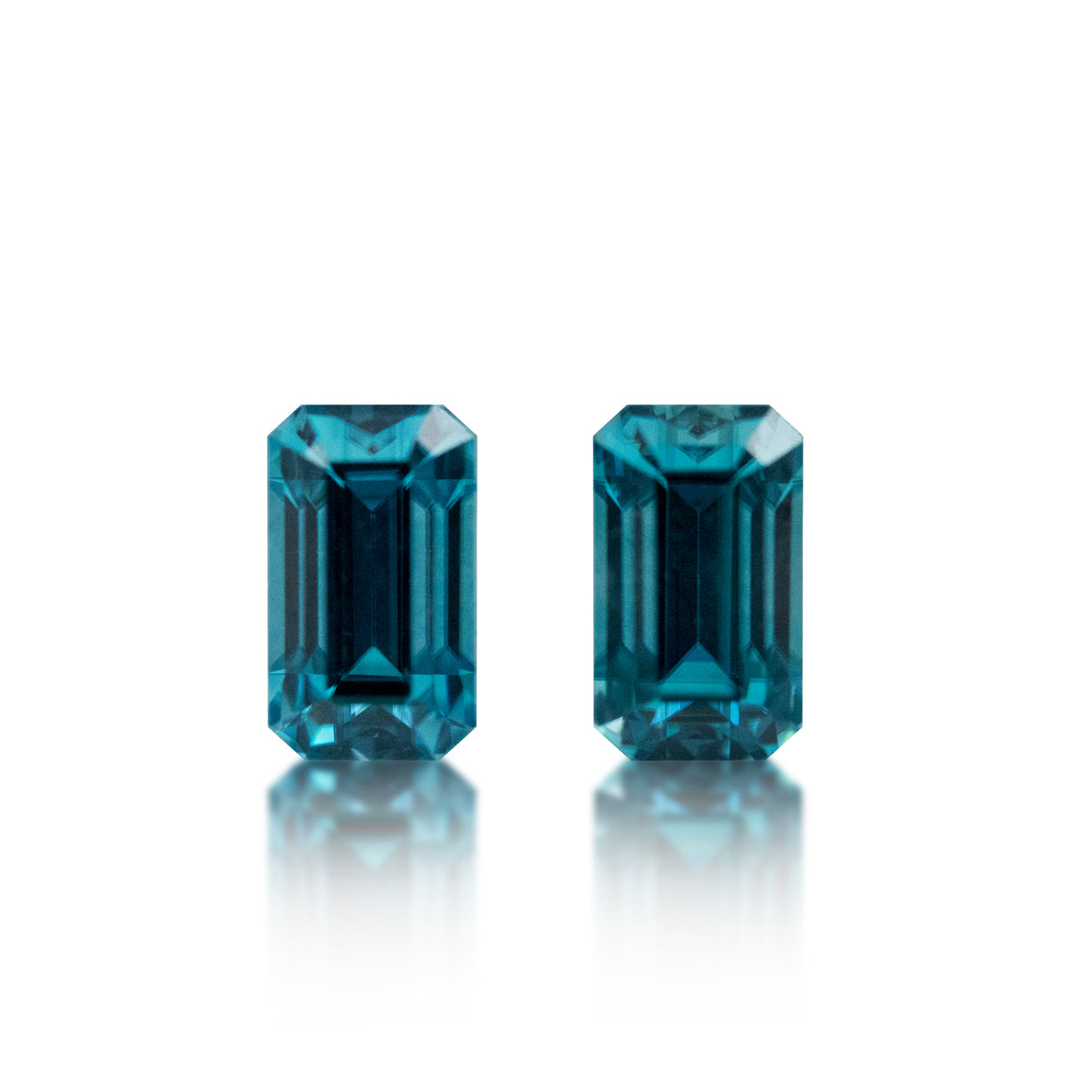 A pair of rectangular emerald-cut blue gemstones with a faceted, step-cut appearance, displayed side-by-side on a reflective white surface against a solid white background.